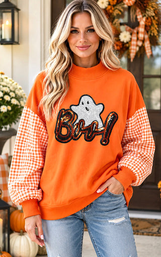 A woman wearing a bright orange crewneck sweatshirt with orange and white gingham checkered sleeves and a sequined 'Boo!' graphic on the front, paired with light blue jeans, standing in a decorated autumn porch setting.