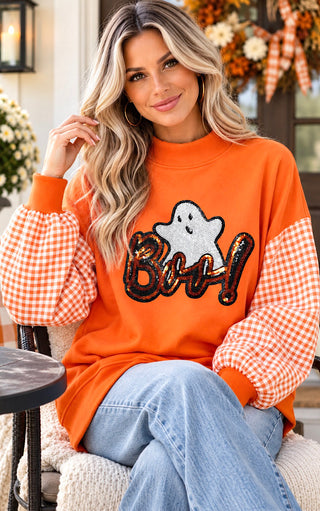 A woman wearing an orange crewneck sweatshirt with orange and white gingham checkered sleeves and a sequined 'Boo!' graphic on the front, paired with light blue jeans.