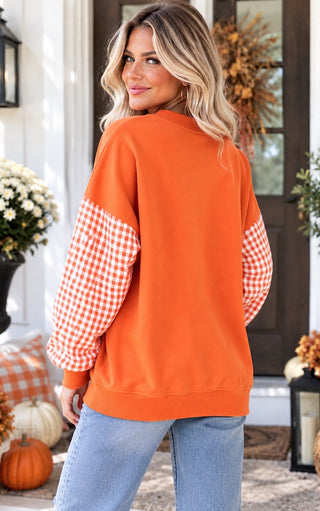 A woman wearing an orange crewneck sweatshirt with orange and white gingham checkered long sleeves, paired with light blue jeans, standing on a decorated autumn porch.