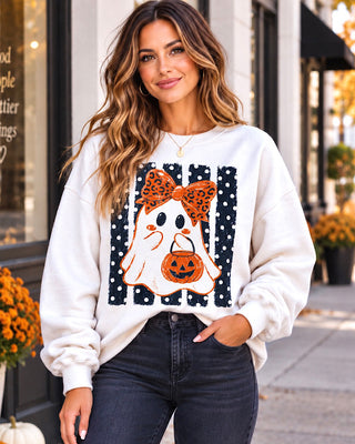 A woman wearing a white crewneck sweatshirt with a Halloween graphic of a ghost, leopard-print bow, and jack-o'-lantern, paired with dark wash jeans, standing on a city sidewalk.