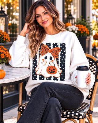 A woman sitting on a patio chair wearing a white crewneck sweatshirt with a Halloween graphic of a ghost, leopard print bow, and pumpkin, holding a coffee cup.