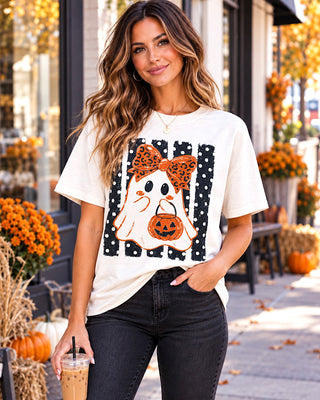 A woman wearing a white graphic t-shirt with a Halloween ghost design, standing outdoors on a sidewalk decorated with pumpkins and fall flowers.