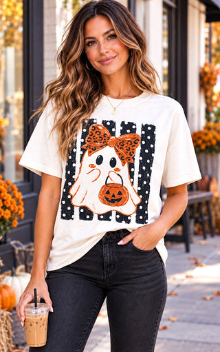 A woman wearing a white graphic t-shirt with a Halloween polka dot ghost design, paired with black jeans, standing outdoors on a fall-themed street.