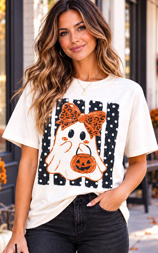 A woman wearing a cream-colored t-shirt with a Halloween graphic featuring a ghost, a jack-o'-lantern, and a leopard-print bow, paired with black jeans.