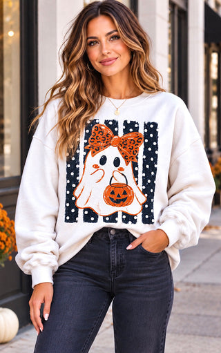 A woman wearing a white crewneck sweatshirt with a large Halloween graphic of a ghost holding a pumpkin, styled with dark wash jeans in an outdoor urban setting.