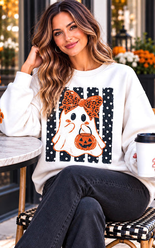 A woman wearing a white crewneck sweatshirt with a Halloween graphic of a ghost in a leopard-print bow holding a jack-o'-lantern on a polka dot background, sitting on a checkered stool and holding a coffee cup.