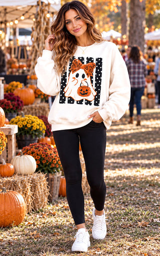 A woman stands outdoors at a fall harvest market wearing a cream-colored crewneck sweatshirt with a Halloween graphic of a ghost in a leopard-print bow holding a pumpkin, paired with black leggings and white sneakers.
