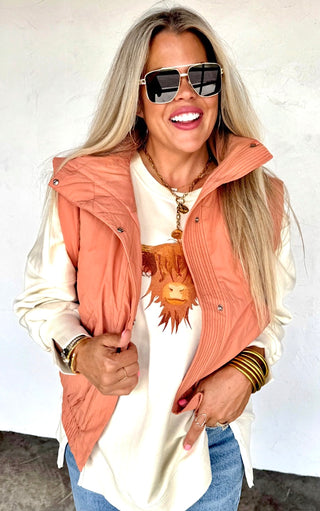 Woman wearing a rust vest over a white shirt with a cow graphic, sunglasses, and gold jewelry.