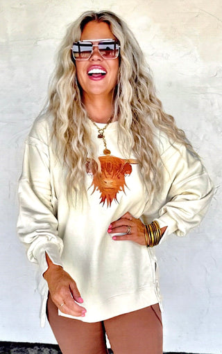Person wearing a cream-colored sweatshirt with a highland cow design, sunglasses, and multiple bracelets on a white background.