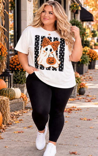 A woman walking outdoors on a fall-themed sidewalk wearing a white graphic t-shirt with a ghost and pumpkin design, black leggings, and white sneakers.