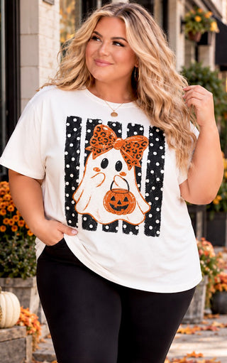 A curvy woman wearing a white graphic t-shirt with a Halloween ghost design, standing outdoors in front of a storefront.