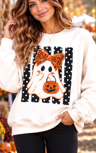 A woman wearing a cream-colored crewneck sweatshirt with a Halloween ghost graphic. The graphic features a white ghost holding an orange jack-o'-lantern pumpkin, adorned with an orange leopard-print bow, set against a background of black and white polka-dot stripes.