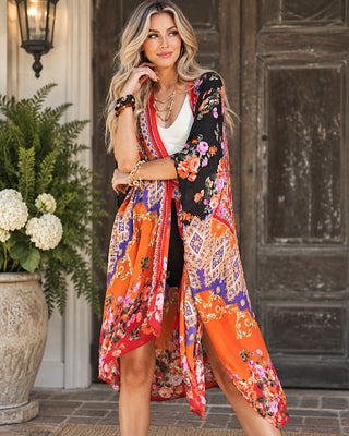 A woman models a multicolored bohemian-style floral kimono cardigan. The garment features a busy pattern of orange, black, red, blue, and pink with paisley and florals, worn over a white top in front of a rustic wooden door.