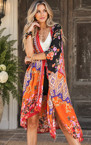 A woman models a vibrant, multicolored bohemian-style floral kimono cardigan. The garment features a busy pattern of orange, black, red, blue, and pink with paisley and floral motifs, worn over a white top in front of a rustic wooden door.