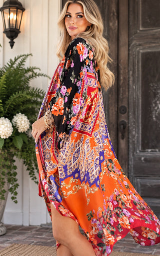 A woman wearing a flowing, long-sleeve kaftan dress with a vibrant multicolor floral and paisley print, standing in front of a rustic wooden door and white wall.