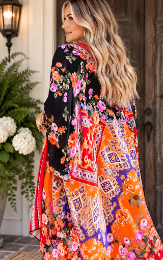 A woman wearing a vibrant, multicolored floral and paisley kaftan-style wrap dress, photographed from behind in an outdoor porch setting.