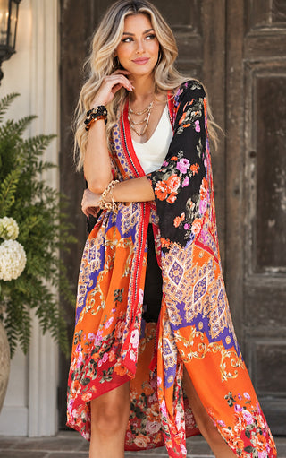 A woman models a vibrant, multicolored floral and geometric print kimono cover-up over a white tank top. The kimono features a black upper section with orange, pink, and purple floral patterns, and a bright orange lower section with purple and gold geometric designs.