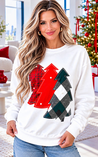 Blonde woman wearing a white crewneck sweatshirt with a graphic of two Christmas trees in red and green plaid patterns, paired with light blue jeans, posed in a cozy holiday living room setting.