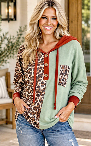 A woman wearing a mixed-media pullover hoodie with a sage green body, leopard print panels, and rust red accents, paired with blue jeans in an outdoor patio setting.