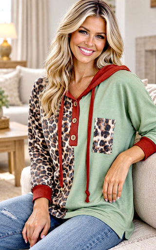 A woman sitting on a couch wearing a mixed-design hoodie with sage green, leopard print, and rust red panels, paired with blue jeans in a cozy living room setting.