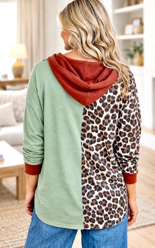 A woman shown from the back wearing a color-blocked hoodie top with a sage green left side, a leopard print right side, and a rust red hood and cuffs, in a brightly lit living room.