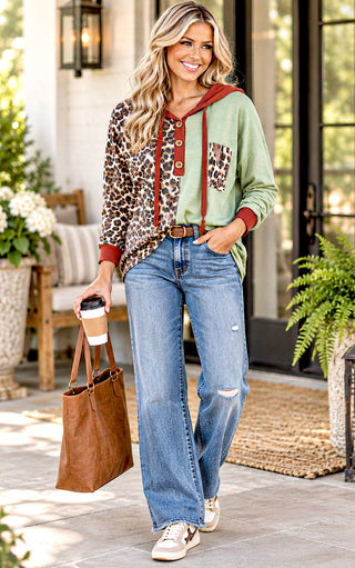 A woman stands on a porch wearing a mixed-print hoodie with leopard print and sage green panels, blue jeans, and white sneakers, while holding a brown tote bag and a coffee cup.