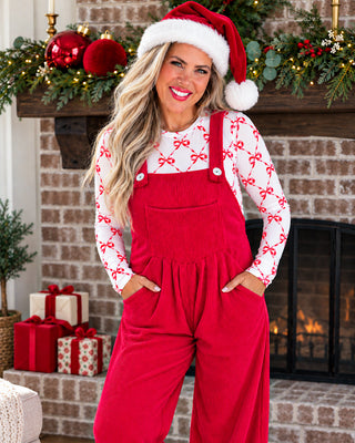 A woman wearing a festive holiday outfit consisting of red corduroy overall jumpsuits over a white long-sleeve shirt with red bow patterns, a red Santa hat, and a matching Santa hat with white trim. She is posing in front of a decorated brick fireplace with Christmas garlands, ornaments, and wrapped gifts.