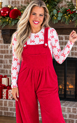 A woman wearing a festive red  overall jumpsuit over a white mesh long-sleeve shirt with a red bow pattern, standing in front of a decorated brick fireplace with holiday garlands and wrapped gifts.