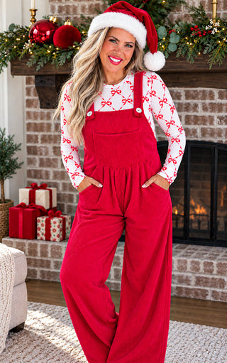 A woman wearing red holiday overalls and a white long-sleeve shirt with red bow patterns, styled with a Santa hat, standing in front of a decorated brick fireplace.