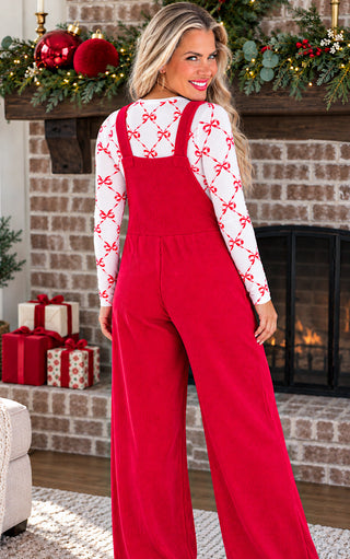 Back view of a woman stands in front of a decorated fireplace wearing a bright red wide-leg jumpsuit over a white long-sleeve shirt with a red bow pattern. The scene includes holiday garlands, ornaments, wrapped gifts, and a lit fireplace.