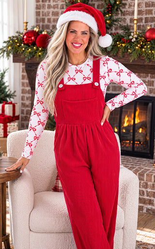 A woman wearing a red ribbed overalls and a white mesh long-sleeve shirt with a red bow pattern, accessorized with a Santa hat, posed in front of a cozy fireplace decorated for Christmas.