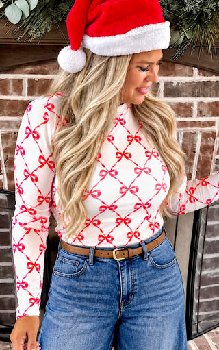 A woman wearing a red and white mesh bow patterned long-sleeve top, blue jeans, and a red Santa hat, standing in front of a brick fireplace.
