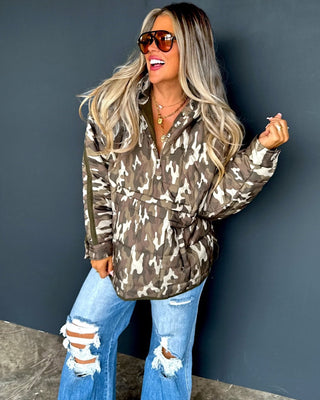 Woman wearing a camouflage puffer jacket and ripped jeans against a dark blue wall.