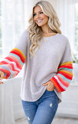 Woman wearing a gray sweater with colorful sleeves and blue jeans indoors.