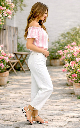 Woman in a pink top and white barrel jeans standing on a stone path with flowers around.