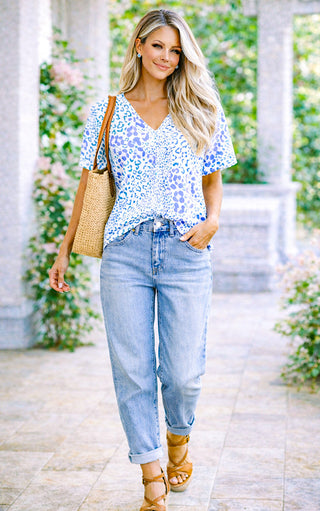 Woman in a blue floral top and jeans walking outdoors with a straw bag.