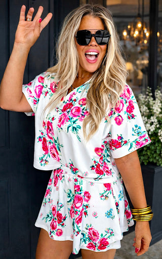 Woman wearing a floral skirt set with sunglasses and a gold bracelet, waving.