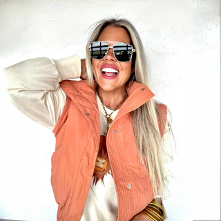 Woman wearing an orange puffy vest and sunglasses against a white background