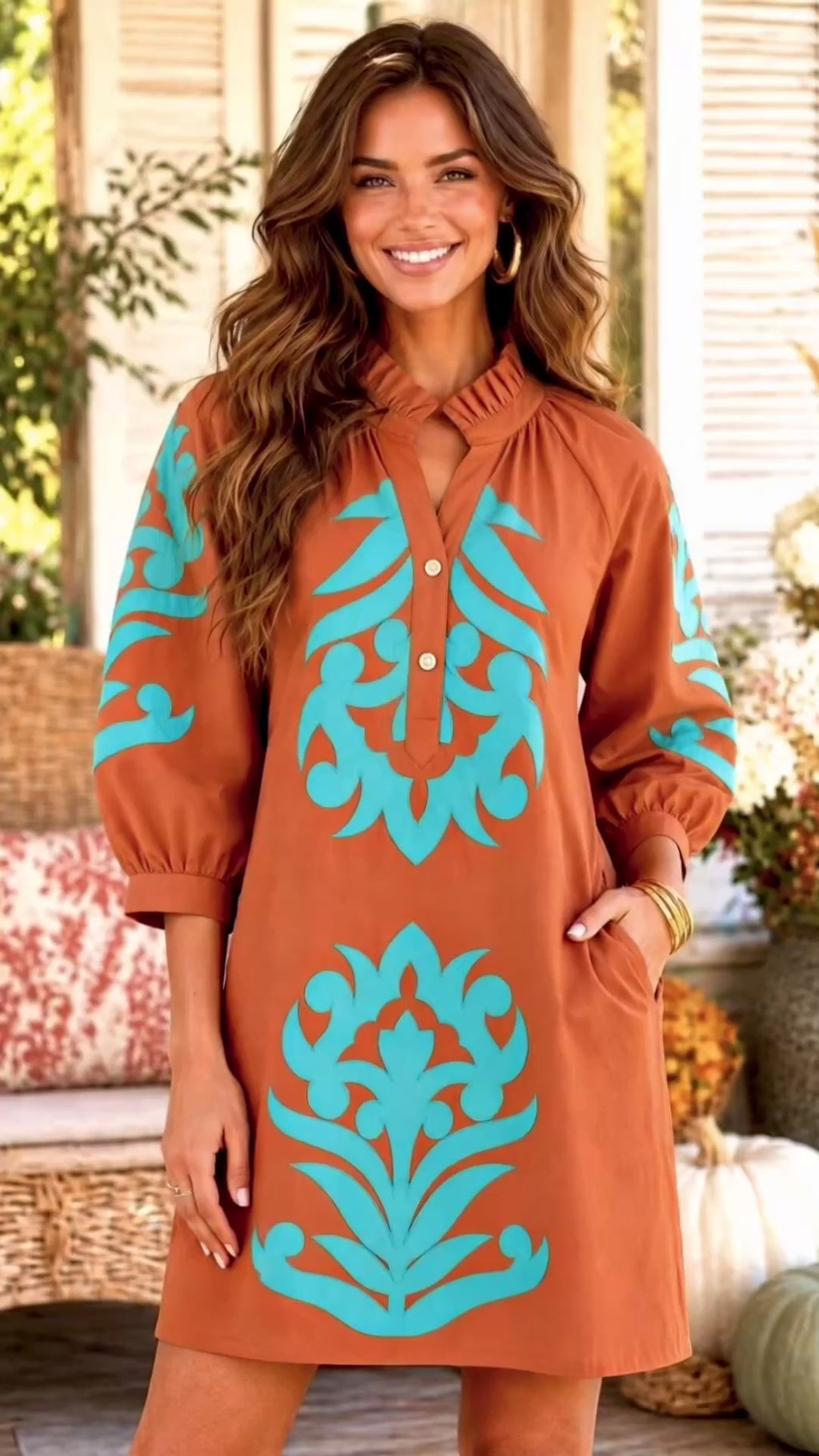 Video of a woman modeling a toffe brown tunic dress with turquoise blue botanical patterns, standing on a porch decorated with pumpkins, flowers, and a wicker chair.