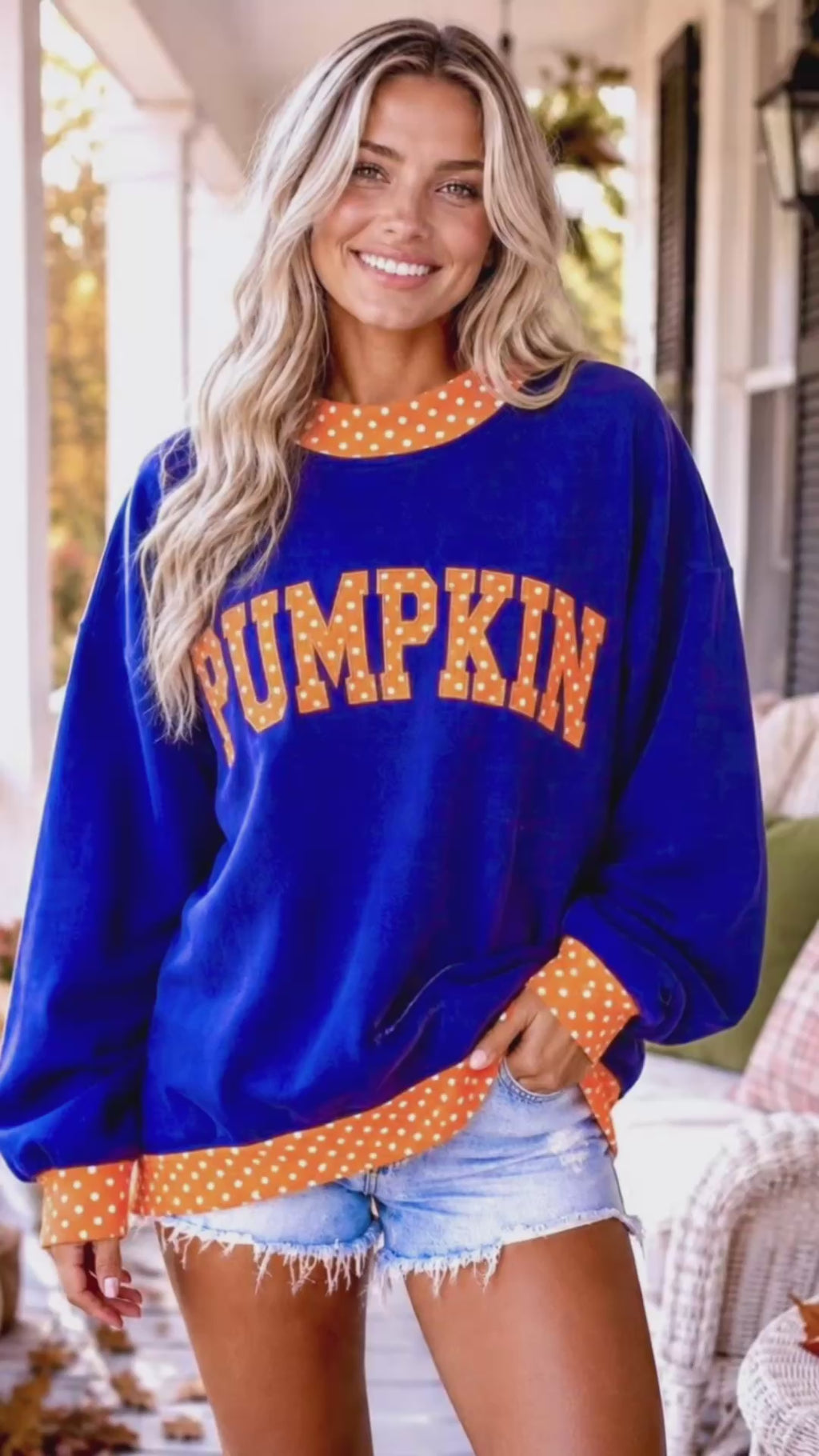 Video of A woman wearing a royal blue long-sleeve sweatshirt with orange and white polka-dot trim and 'PUMPKIN' printed across the front in orange letters, paired with blue jeans in an outdoor porch setting.