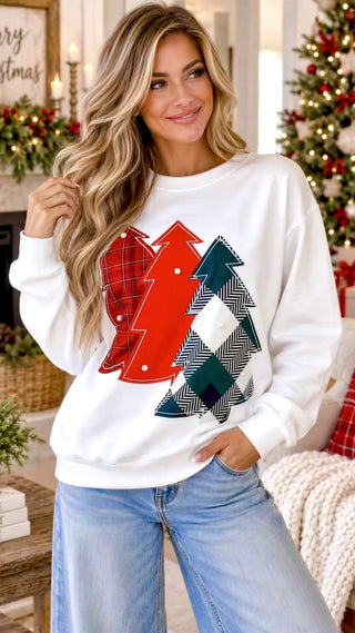 Video of A woman wearing a white crewneck sweatshirt with a graphic of three Christmas trees in red, green, and black-and-white herringbone patterns, posed in a cozy holiday living room setting.