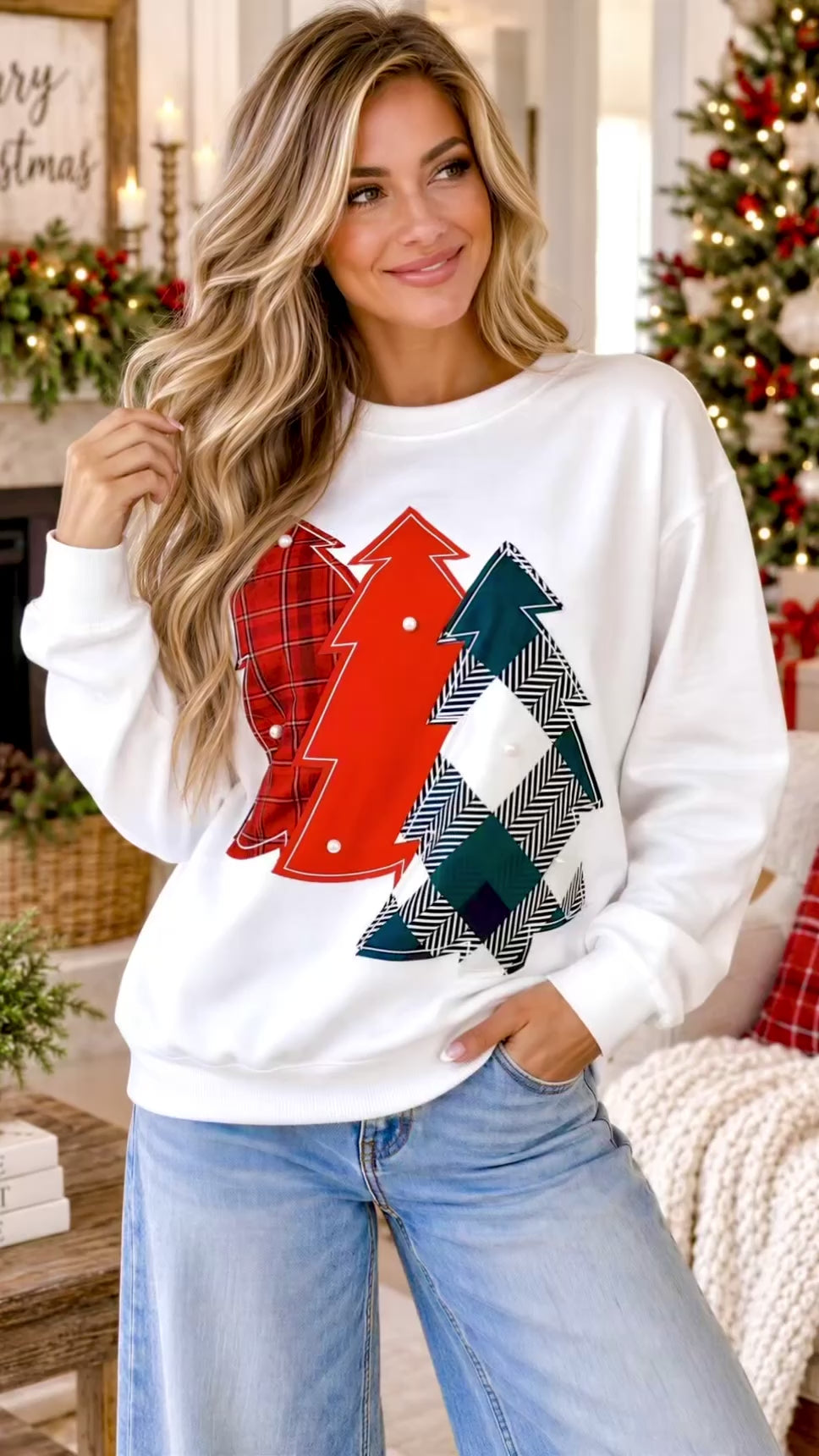 Video of A woman wearing a white crewneck sweatshirt with a graphic of three Christmas trees in red, green, and black-and-white herringbone patterns, posed in a cozy holiday living room setting.