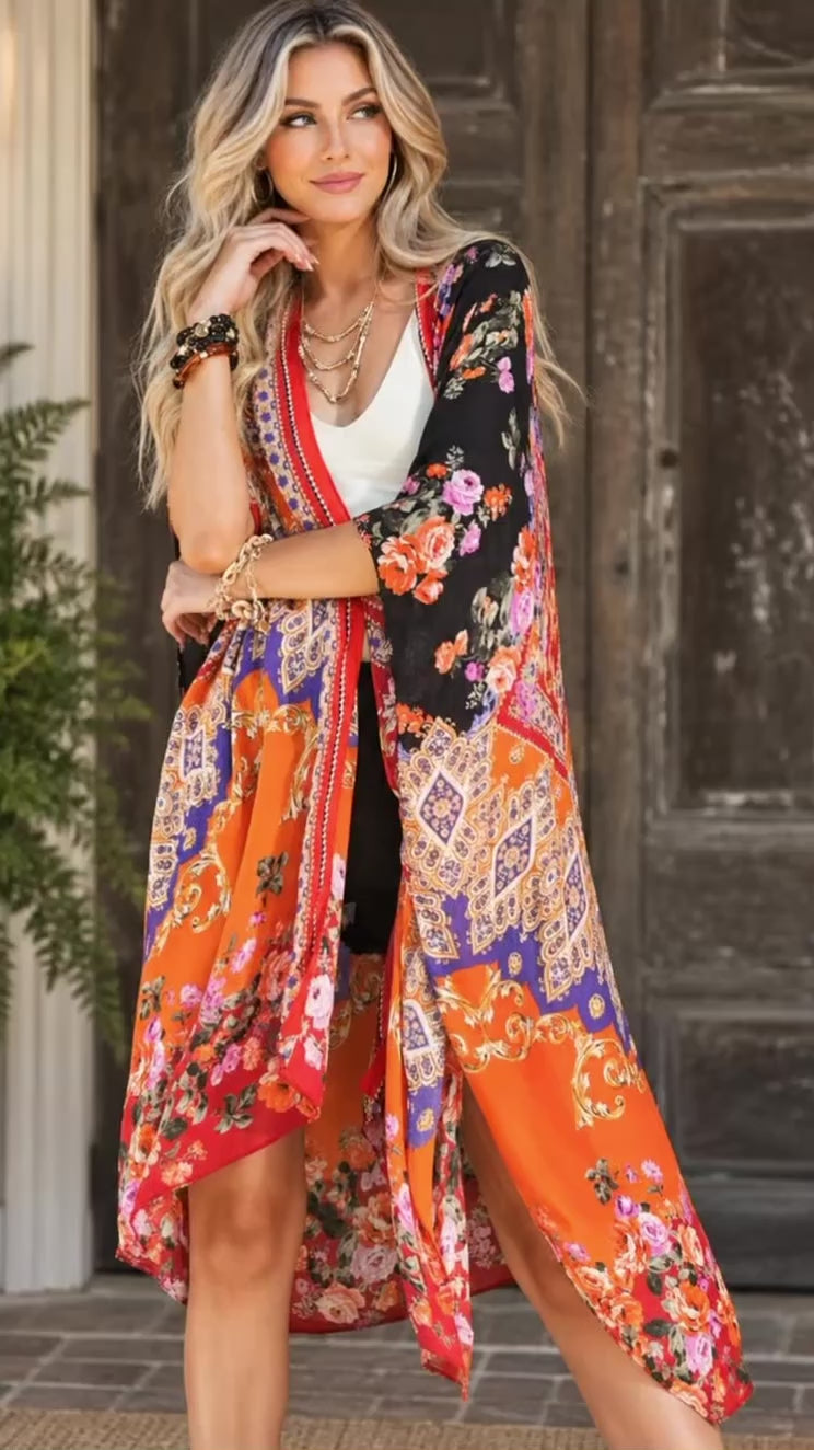 Video of a woman modeling a vibrant, multicolored bohemian-style floral kimono cardigan. The garment features a busy pattern of orange, black, red, blue, and pink with paisley and floral motifs, worn over a white top in front of a rustic wooden door.