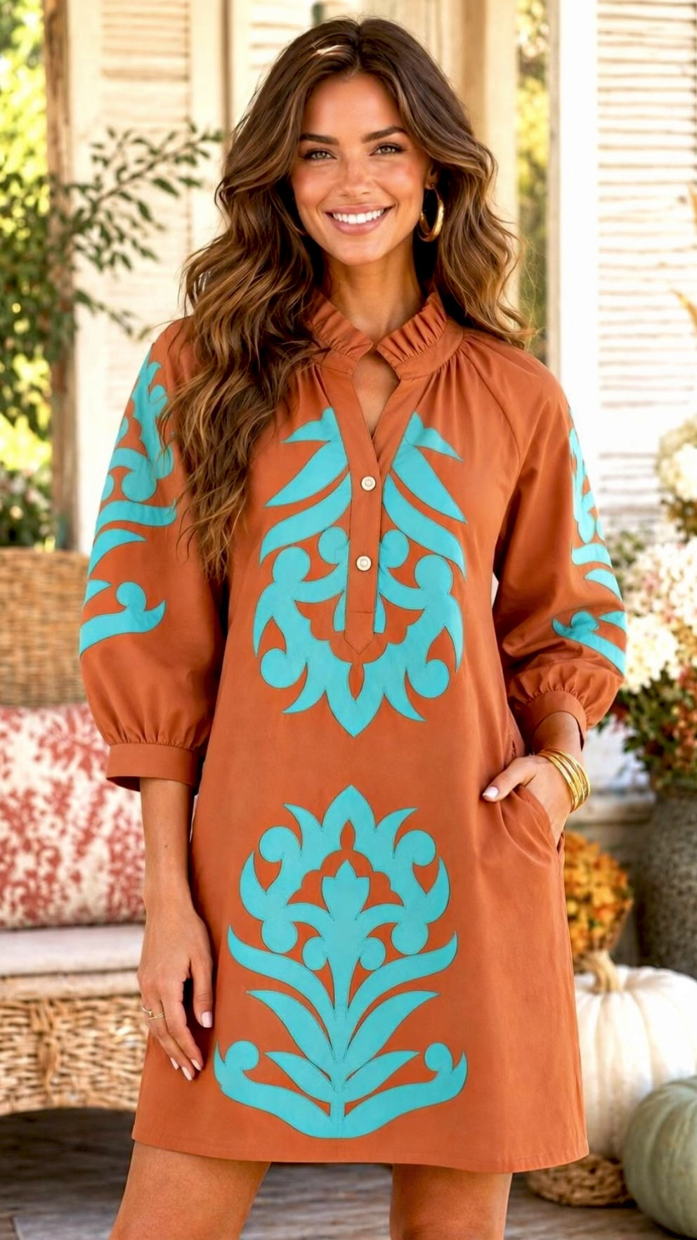 Video of a woman modeling a rust brown  shift dress with turquoise damask-style patterns on the sleeves and front, set against a warm, autumnal porch background with pumpkins and wicker.
