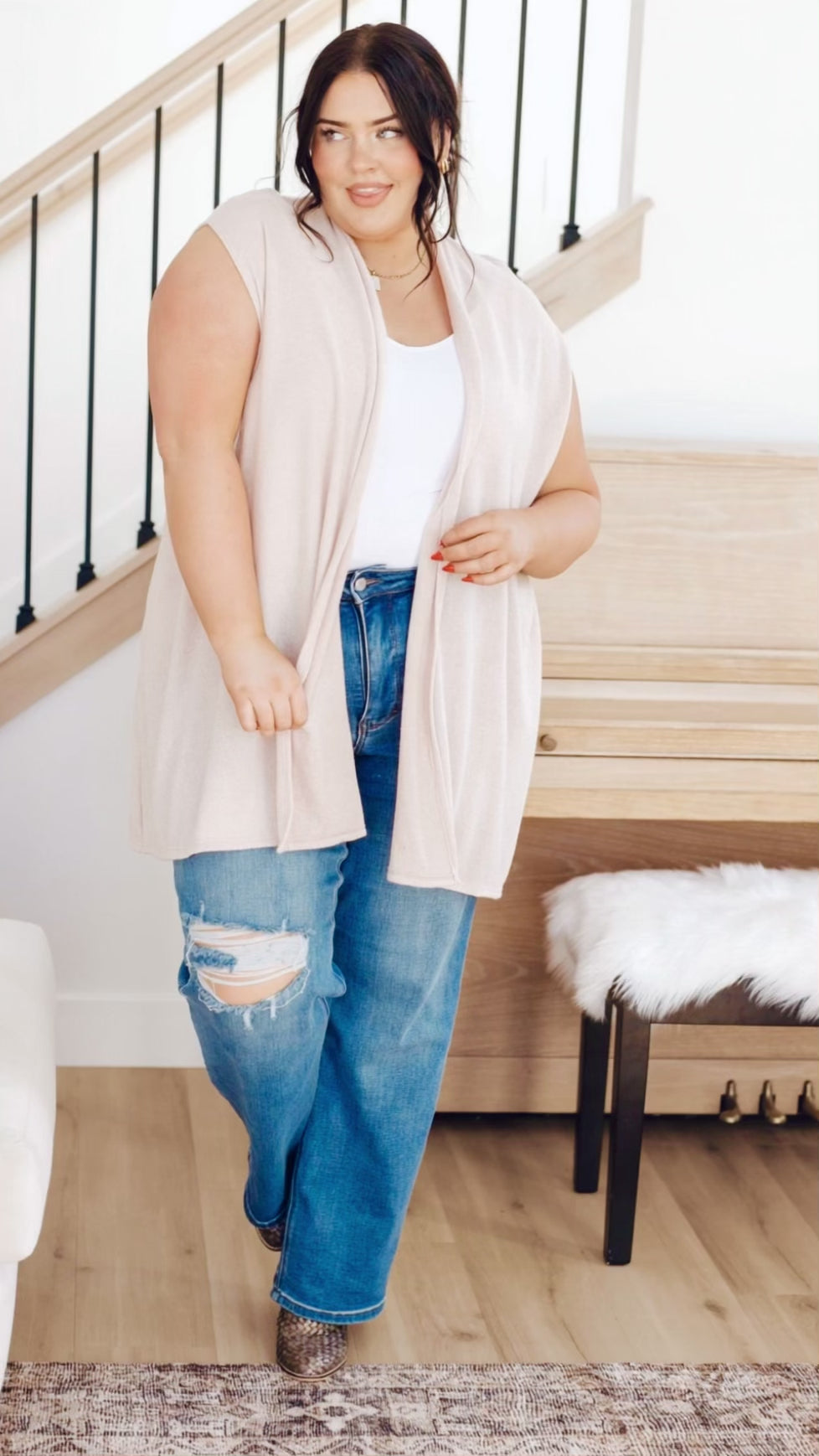 Plus-size model wearing cream sleeveless cardigan with pockets, styled over a white tank and distressed jeans, moving naturally to show fit.
