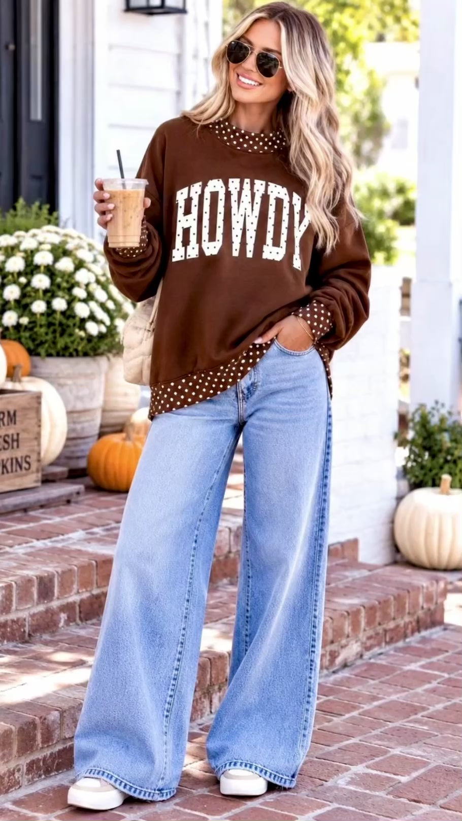 Video of A woman wearing a brown crewneck sweatshirt with 'HOWDY' printed in white across the front, featuring white polka-dot trim at the collar, cuffs, and hem. She is wearing light blue jeans, sunglasses, and holding an iced coffee in an outdoor setting.