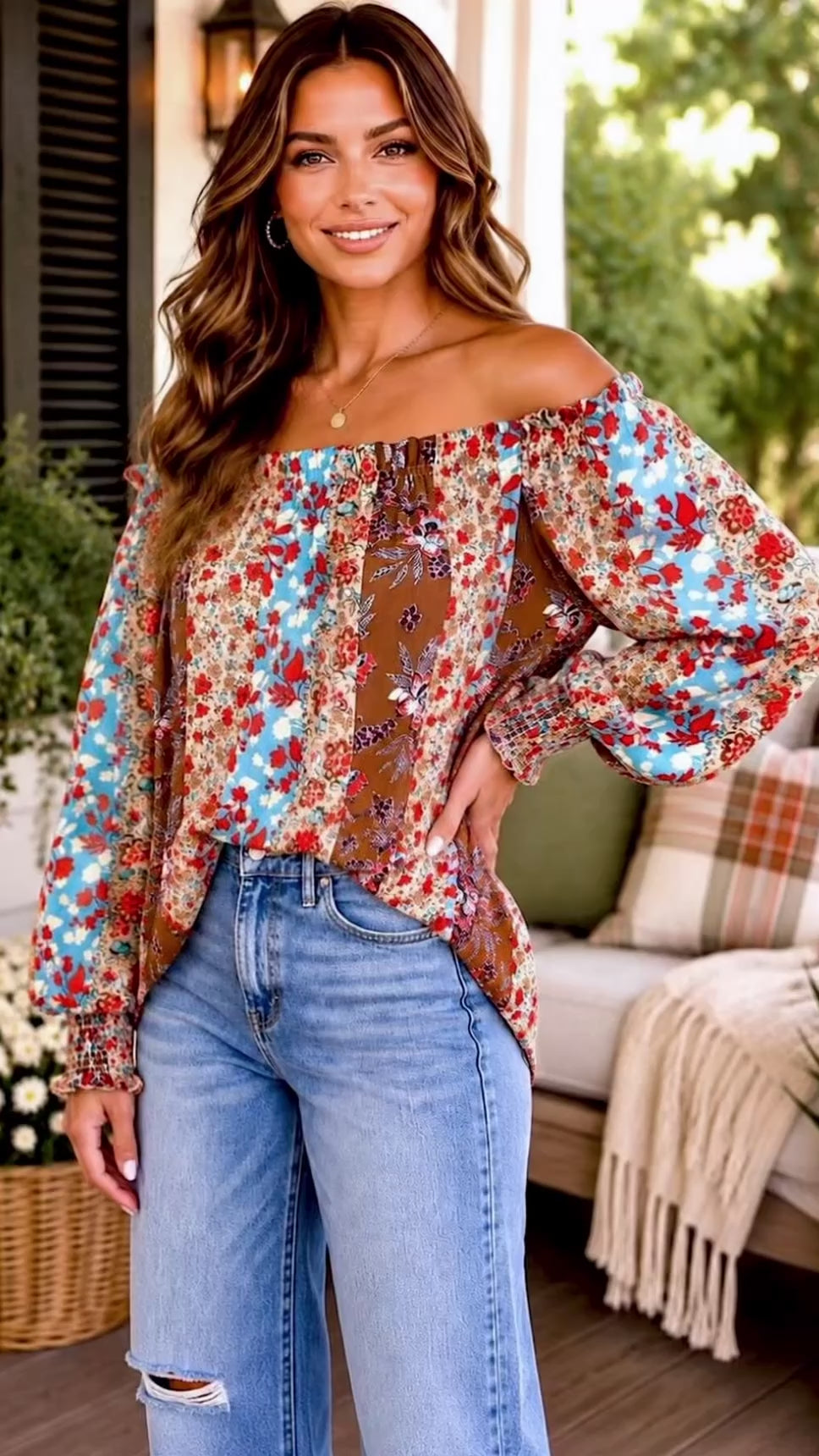 Video of a woman wearing a colorful off-the-shoulder long-sleeve blouse with a red, turquoise, and  brown floral pattern, paired with blue jeans, standing in a bright, sunny porch setting.