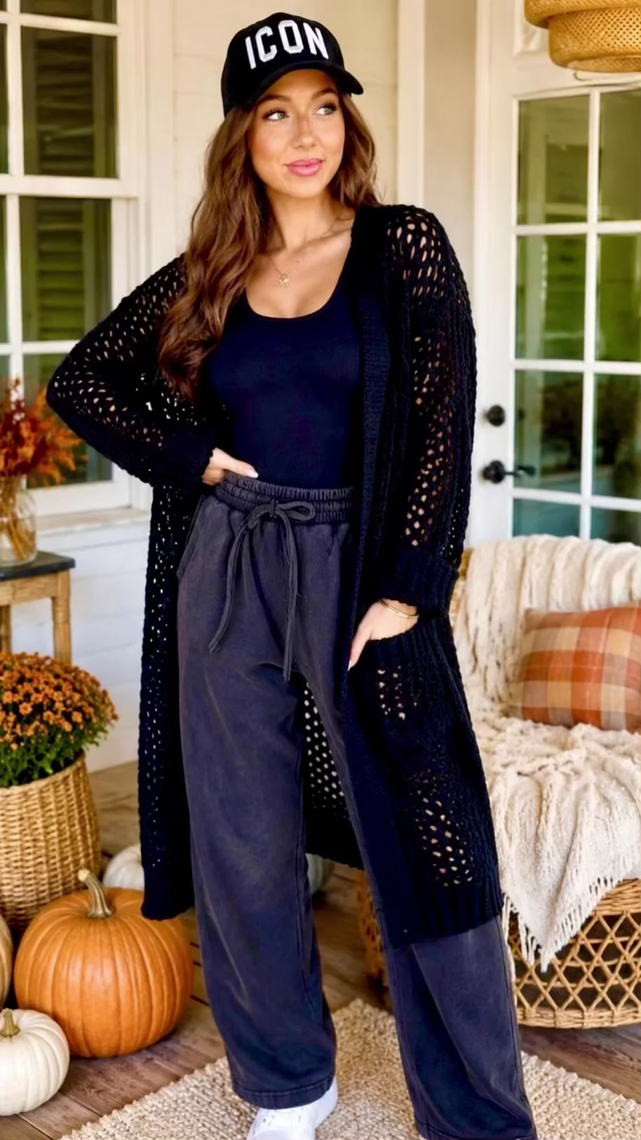 Video of a woman wearing standing on a porch wearing a long black open-knit cardigan over a black top and dark gray drawstring pants, with a black cap that reads 'ICON'. Pumpkins, flowers, and a porch chair are visible in the background.