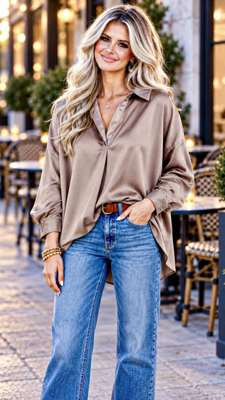 Video of a woman posing in a stylish outdoor cafe setting, wearing an oversized brown satin button-down shirt, blue wide-leg jeans, and carrying a brown leather handbag